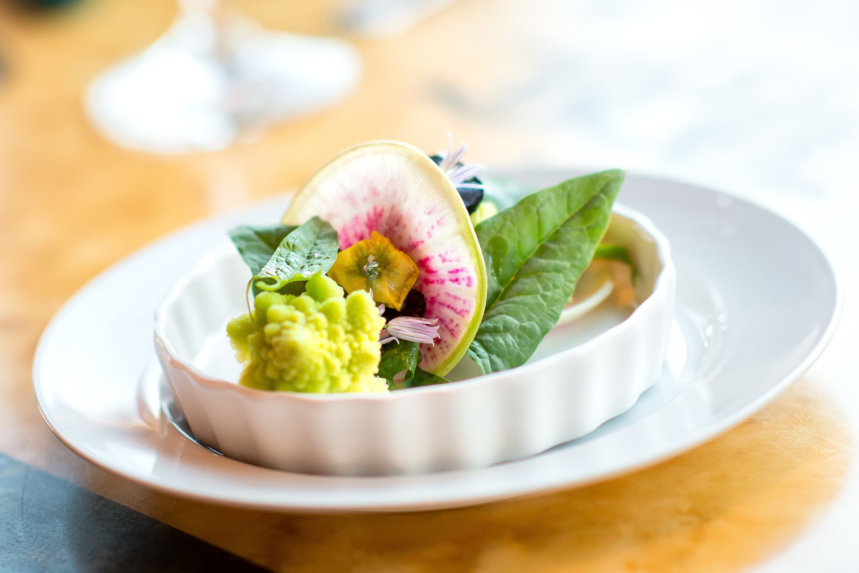 Een witte schaal met een groene salade, een plak radijs en een plak kiwi op een wit bord.