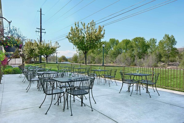 Une terrasse extérieure avec des tables et des chaises en métal, entourée d’une clôture verte et d’arbres.