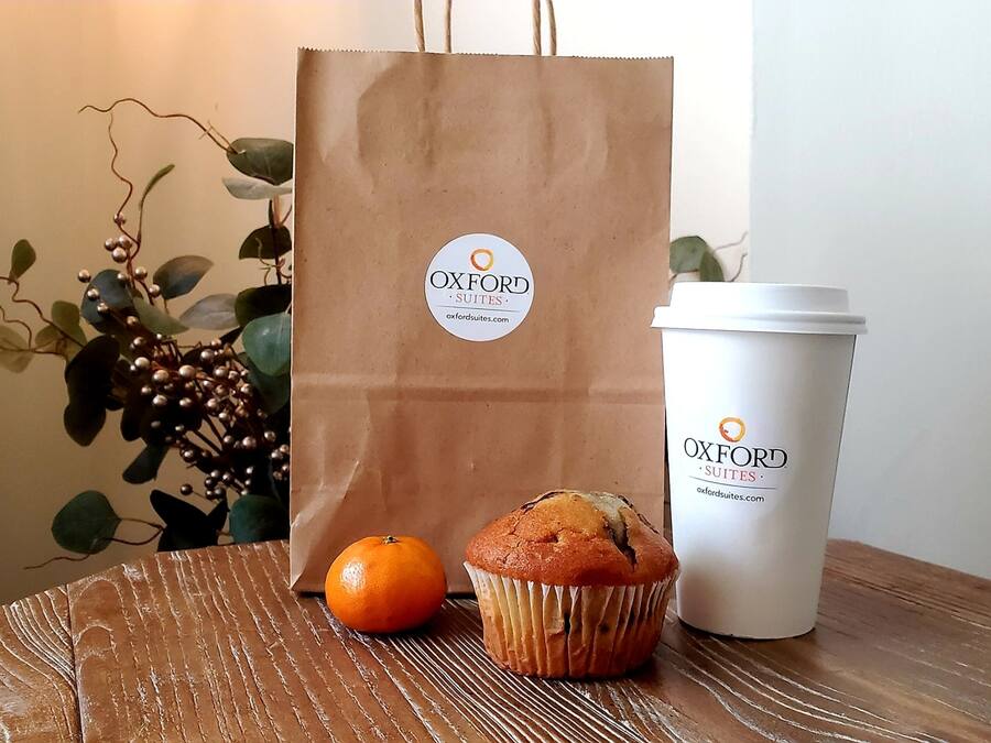 A brown paper bag and a white cup with the Oxford Suites logo, a muffin, and an orange on a wooden table.