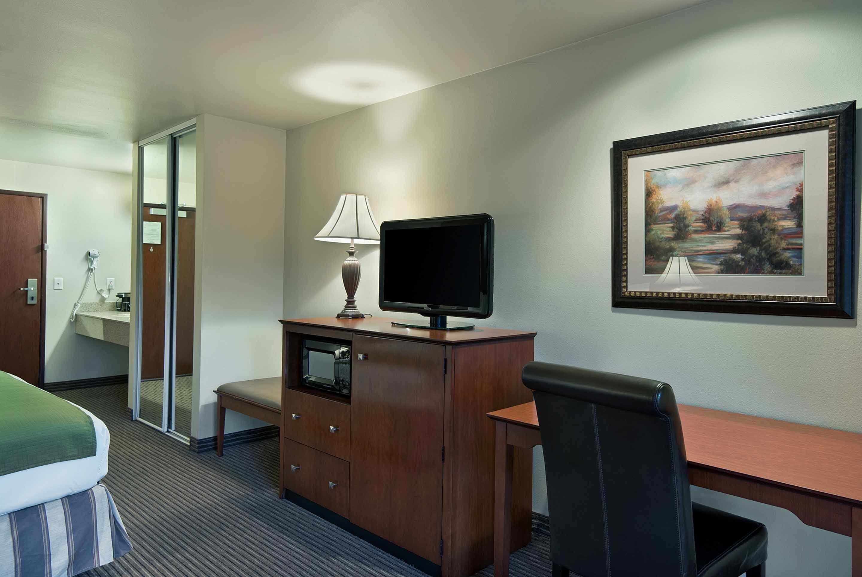 A hotel room with a bed, a desk with a computer, a chair, a TV, a lamp, a framed picture on the wall, and a bathroom with a mirror and sink.