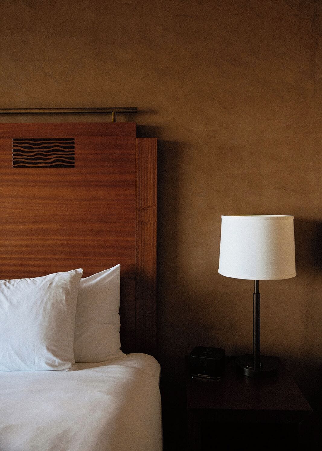 A hotel room with a wooden headboard, white bedding, and a bedside table with a lamp.