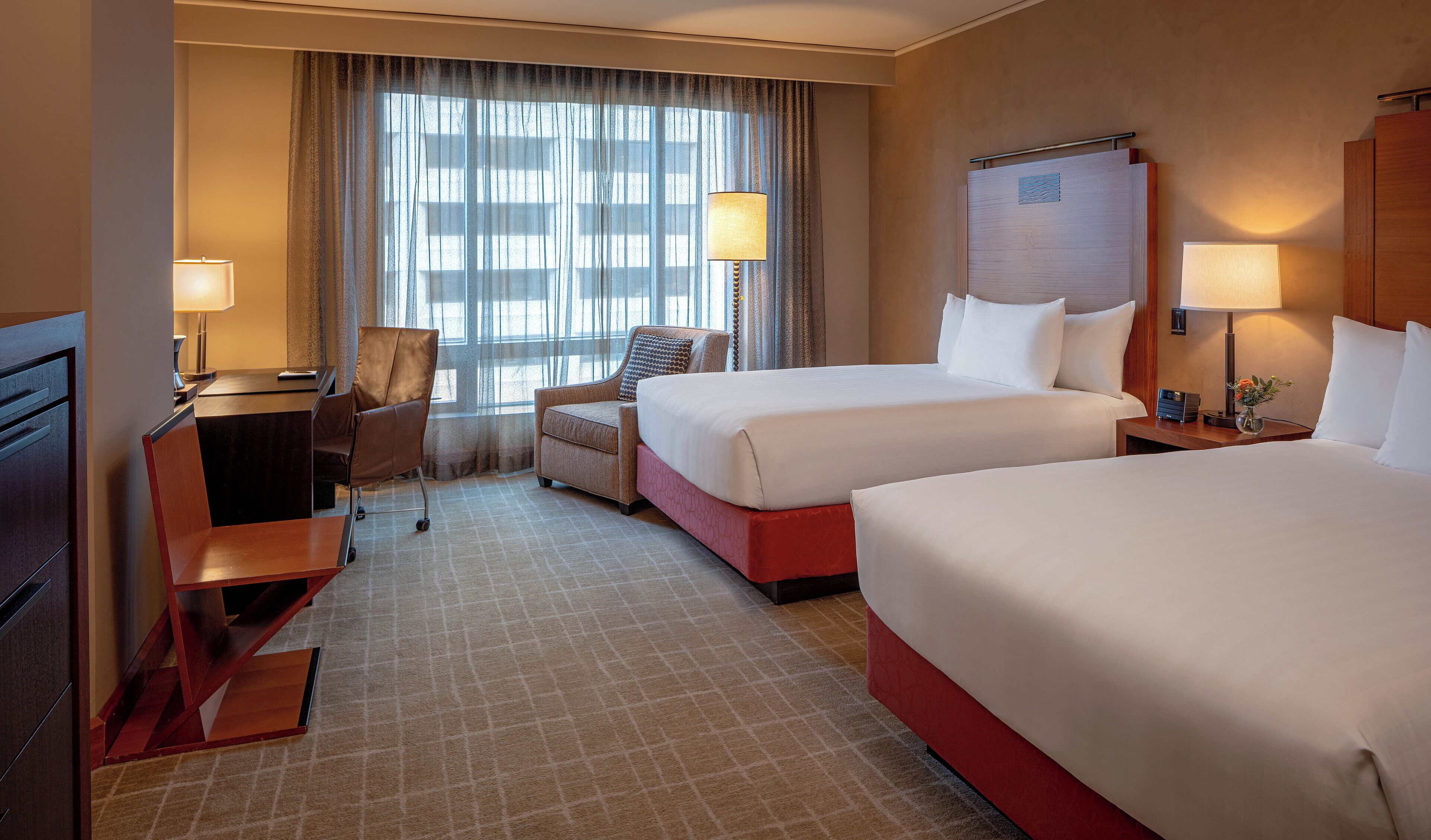 Ein Hotelzimmer mit zwei Betten, einem Schreibtisch, einem Stuhl und einem großen Fenster mit Vorhängen.