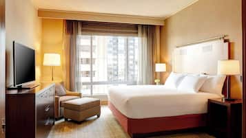 A hotel room with a large bed, a television, a desk with a lamp, and a view of the city through the window.