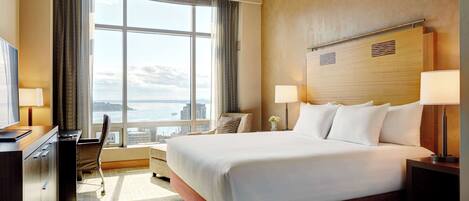 A hotel room with a large bed, a desk, a chair, and a view of the city through a large window.