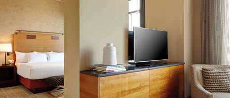 Habitación de hotel con cama, televisor en mueble de madera, una silla y ventana con cortinas.