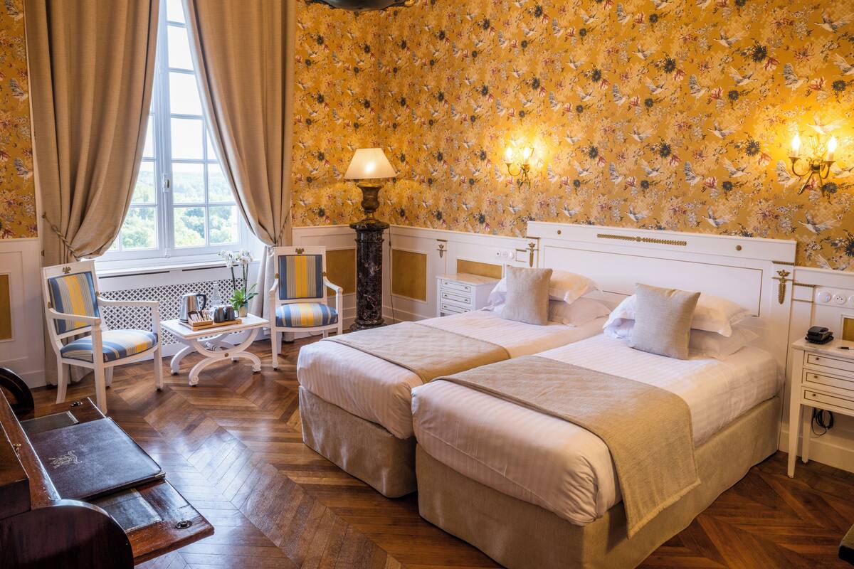 A hotel room with two beds, a sitting area with a chair and a table, a window with curtains, and floral wallpaper.