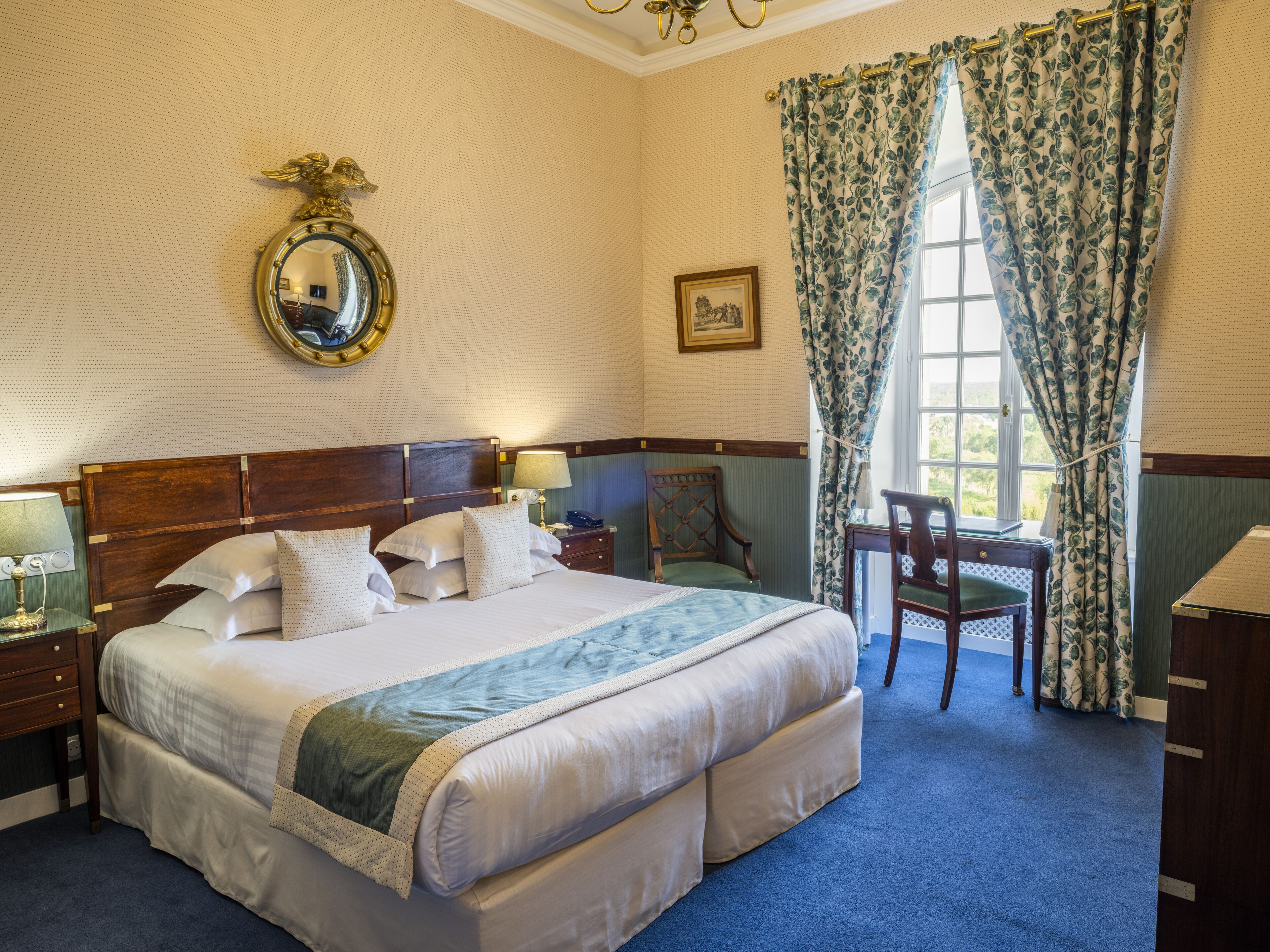 Quarto de hotel com uma cama grande, uma escrivaninha, uma cadeira, um espelho e uma janela com cortinas.