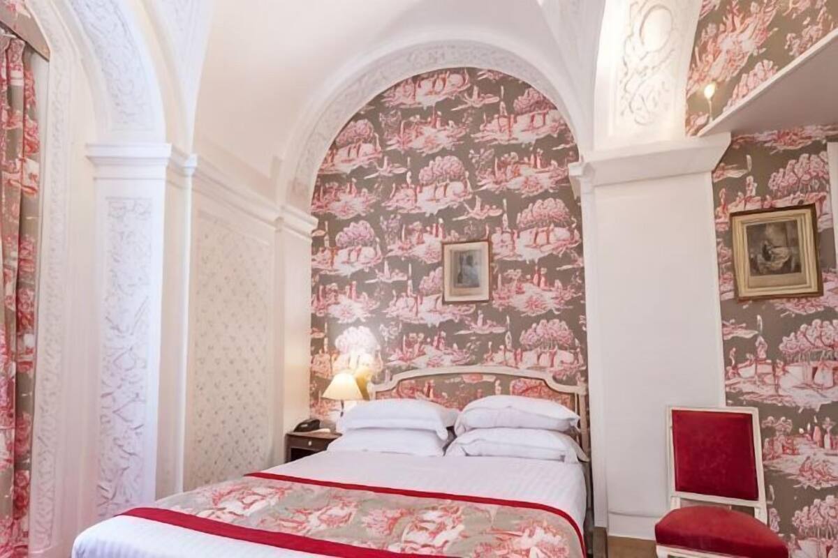 A bedroom with a large bed, a nightstand, a red chair, and floral wallpaper.