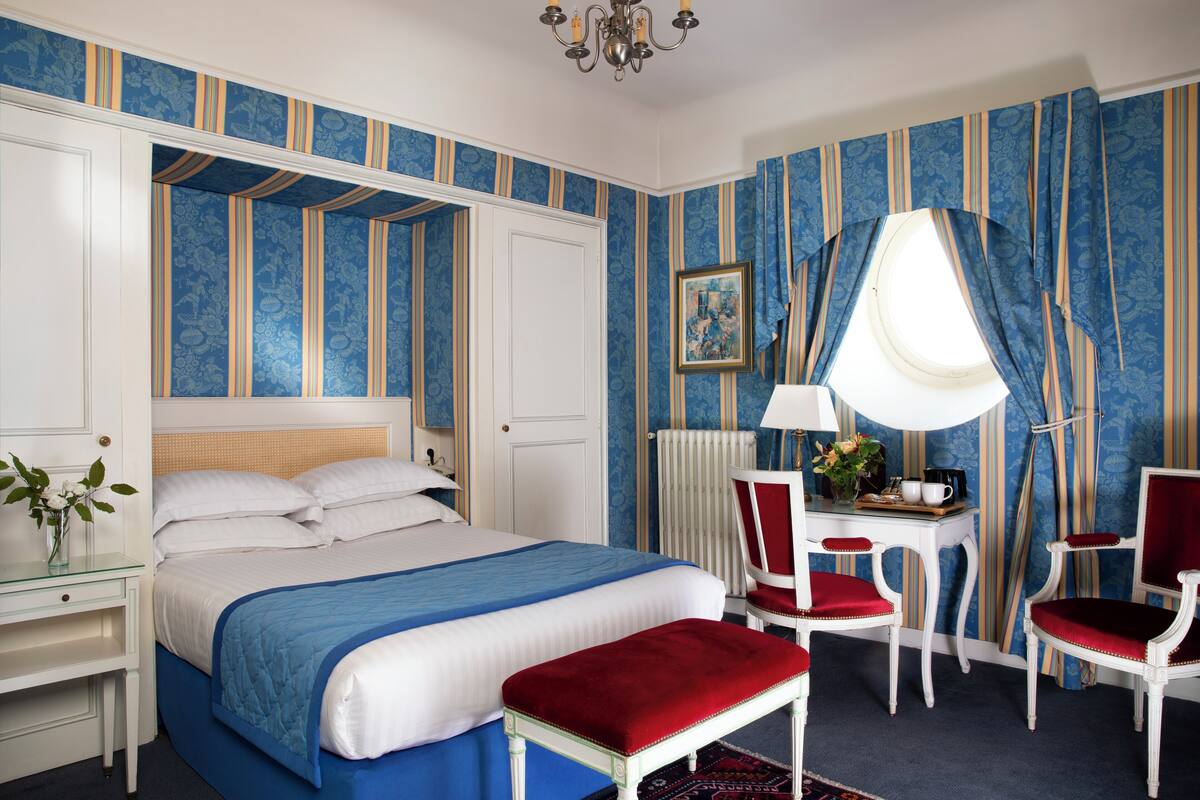 A bedroom with a large bed, a desk with a chair, a window with curtains, and a radiator.
