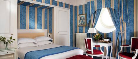 A bedroom with a large bed, a desk with a chair, a window with curtains, and a radiator.