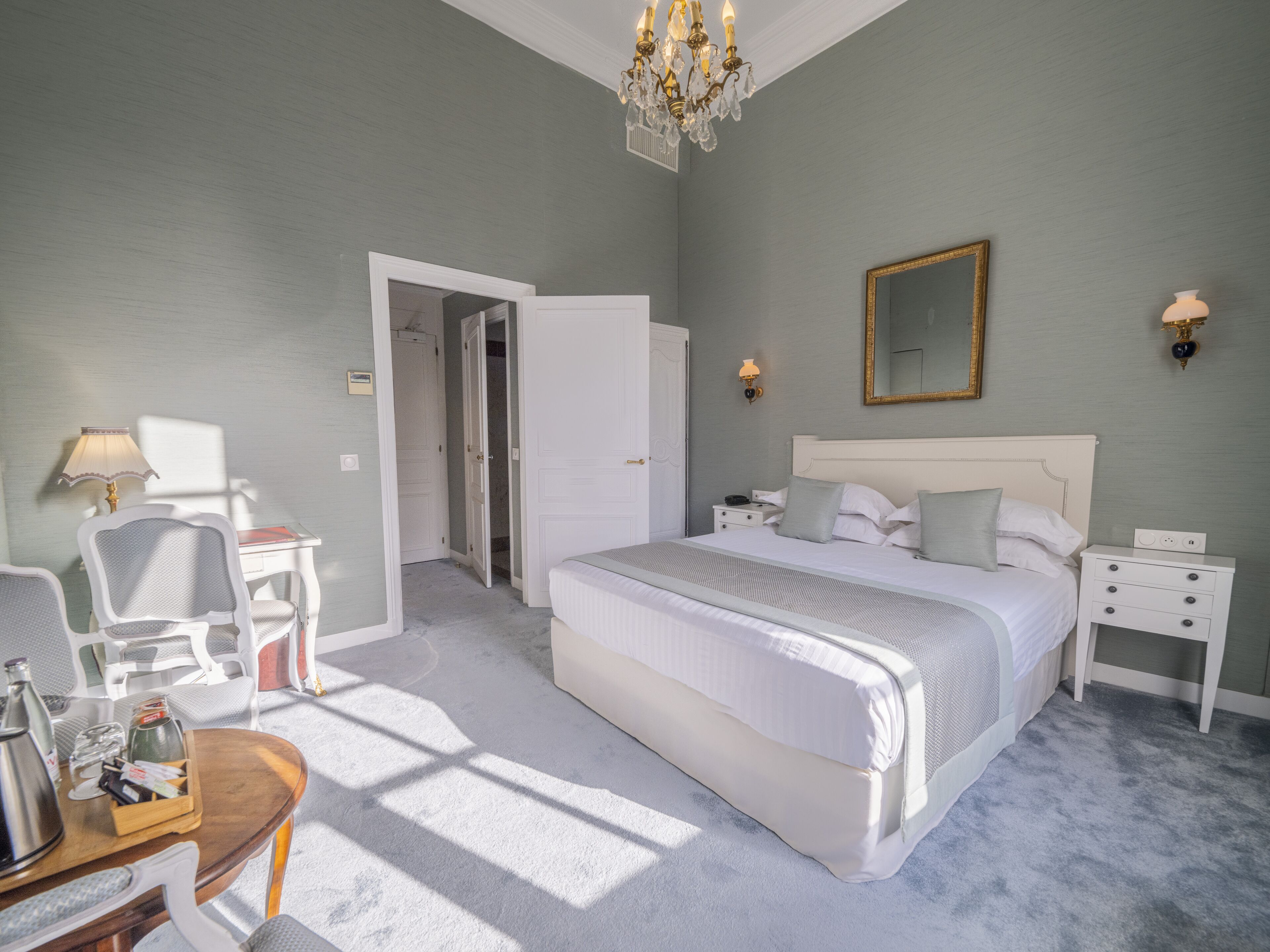 A bedroom with a large bed, bedside tables, a chair, a desk, and a chandelier.