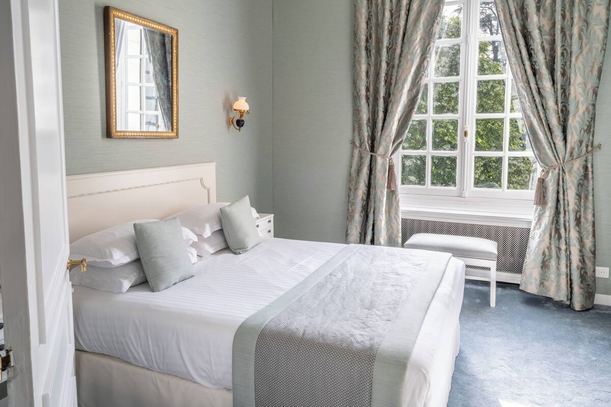A bedroom with a large bed, a window with curtains, a wall-mounted mirror, and a bedside lamp.
