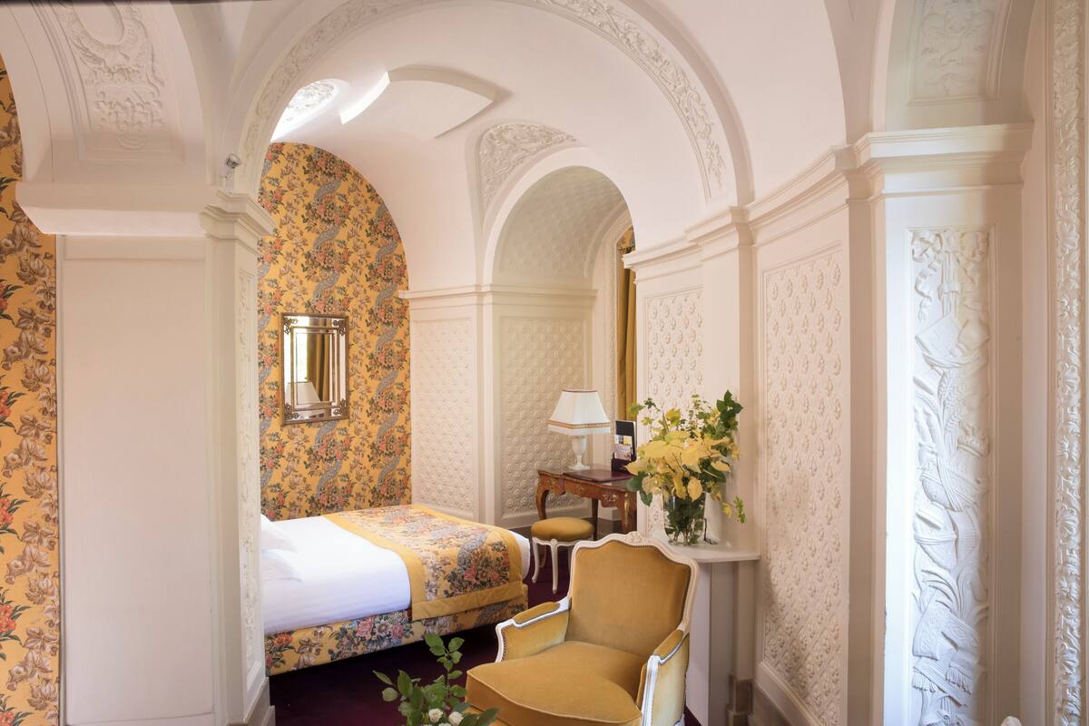 A hotel room with a floral wallpaper, a bed, a chair, and a small table with a lamp.