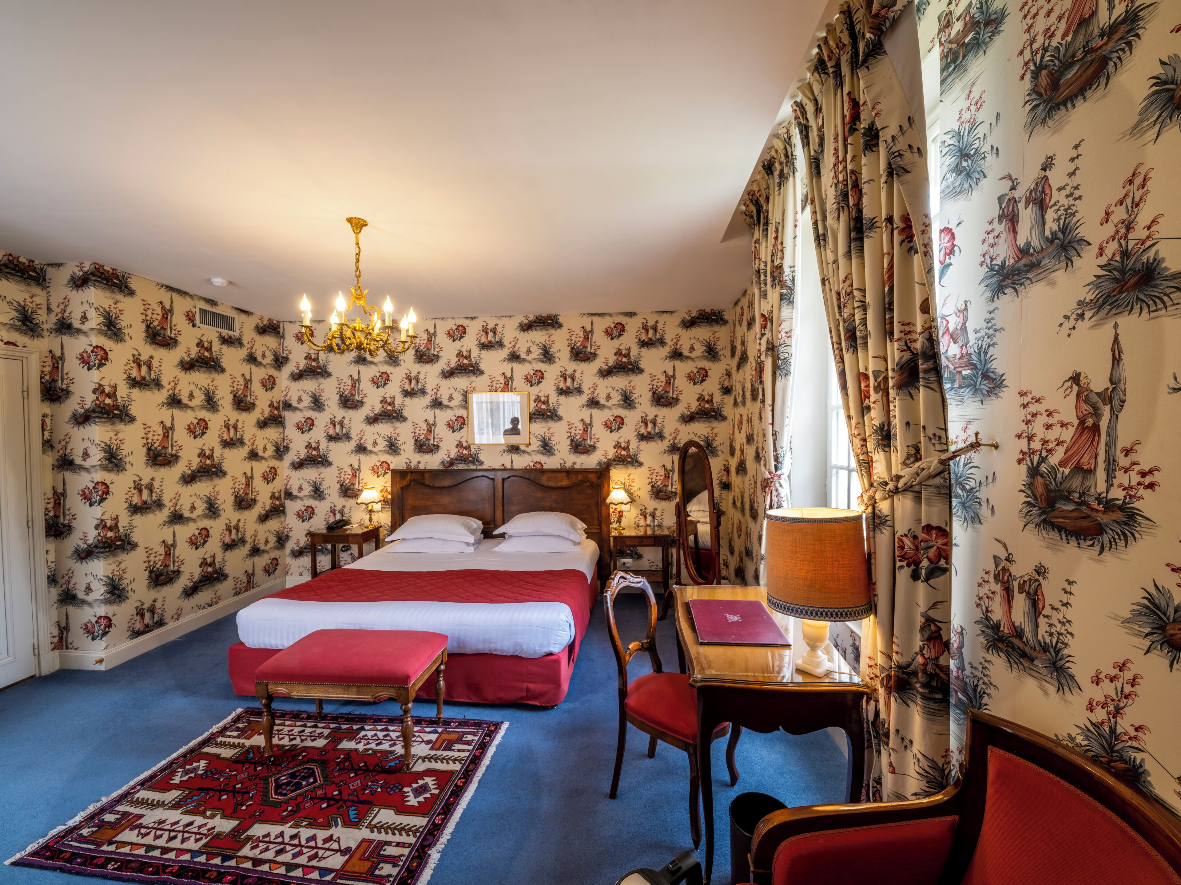 Habitación de hotel con una cama grande, una mesita de noche de madera, un escritorio con lámpara, una silla y una alfombra estampada.