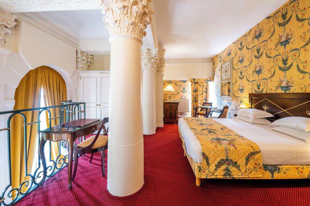 A hotel room with a large bed, a desk with a chair, a dining area with a table and chairs, and a decorative column.