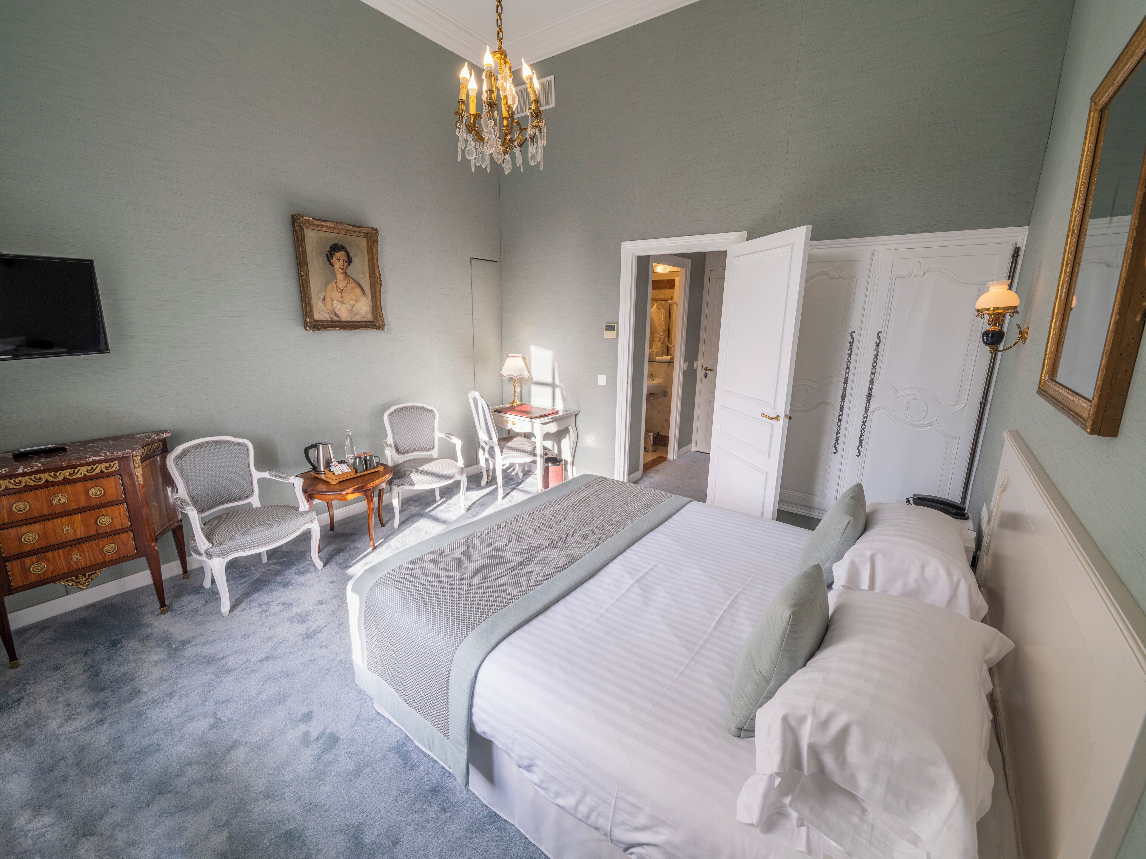 A bedroom with a large bed, a desk with a chair, a dresser, a television, and a chandelier.