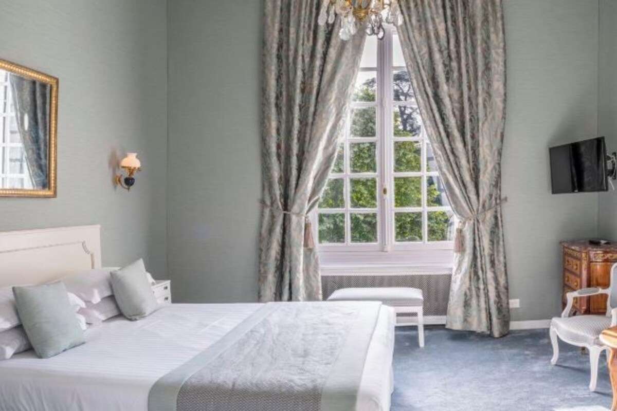 A bedroom with a bed, a chair, a dresser, a television, and a window with curtains.