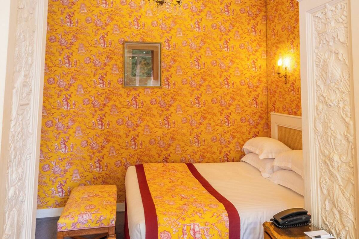 A bedroom with a floral wallpaper, a bed with a red and yellow bedspread, a wooden bench, a nightstand, and a chandelier.
