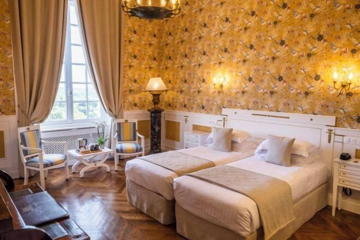 A hotel room with two beds, a sitting area with a chair and table, a lamp, and a window with curtains.