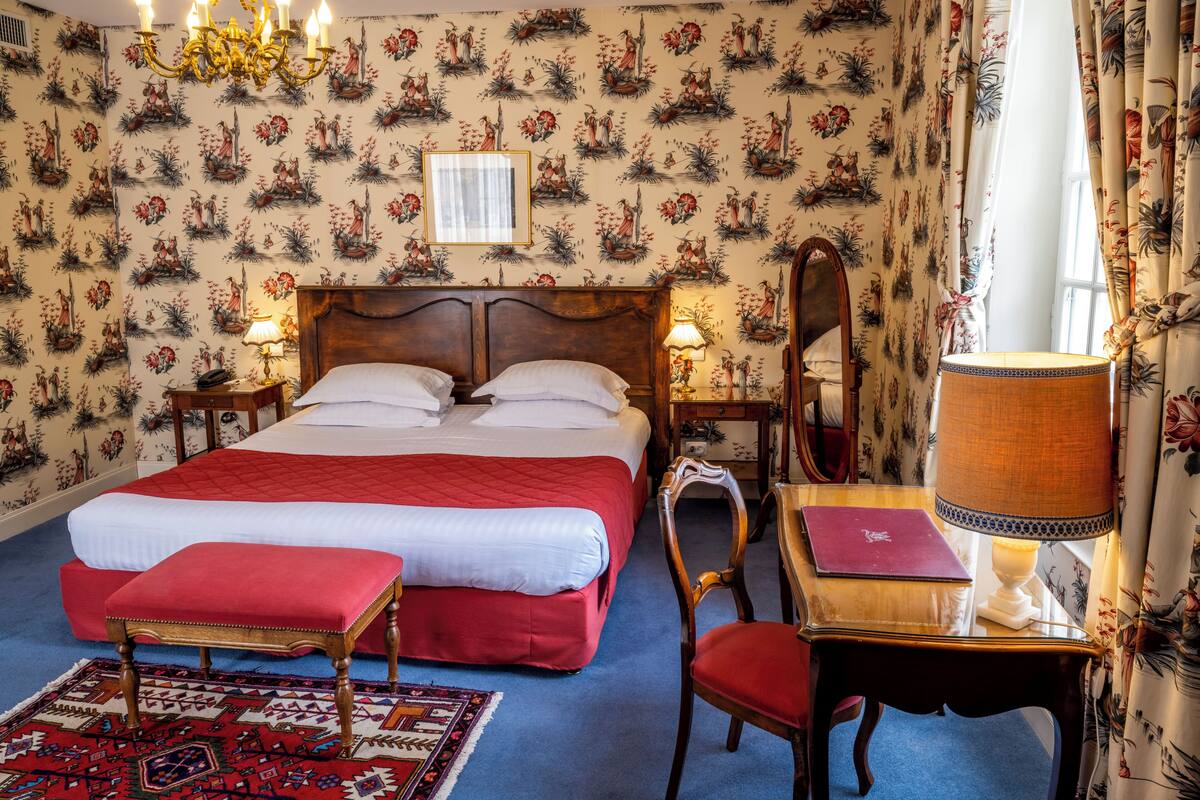 A bedroom with a bed, bedside tables, a chair, a desk, a lamp, a mirror, a chandelier, and floral wallpaper.
