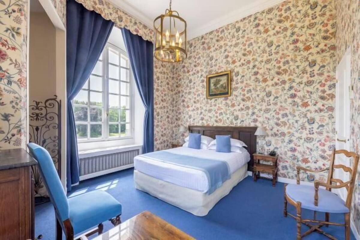 A bedroom with a large bed, a chair, a table, and a window with curtains.