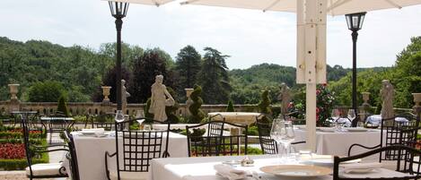An outdoor dining area with tables set for two, surrounded by white umbrellas, with a scenic view of a forested area in the background.