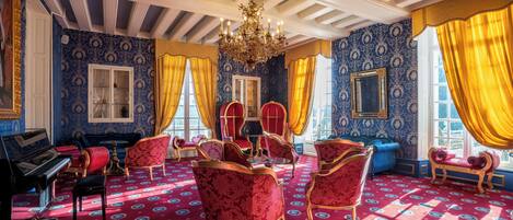 A room with a patterned carpet, a grand piano, red and gold upholstered chairs, and a chandelier.
