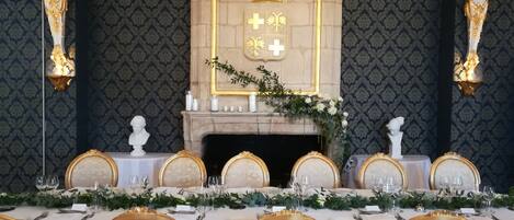 Une salle à manger élégante, avec une longue table dressée pour un repas, entourée de chaises richement décorées, d’une cheminée ornementale et d’un papier peint à motifs.