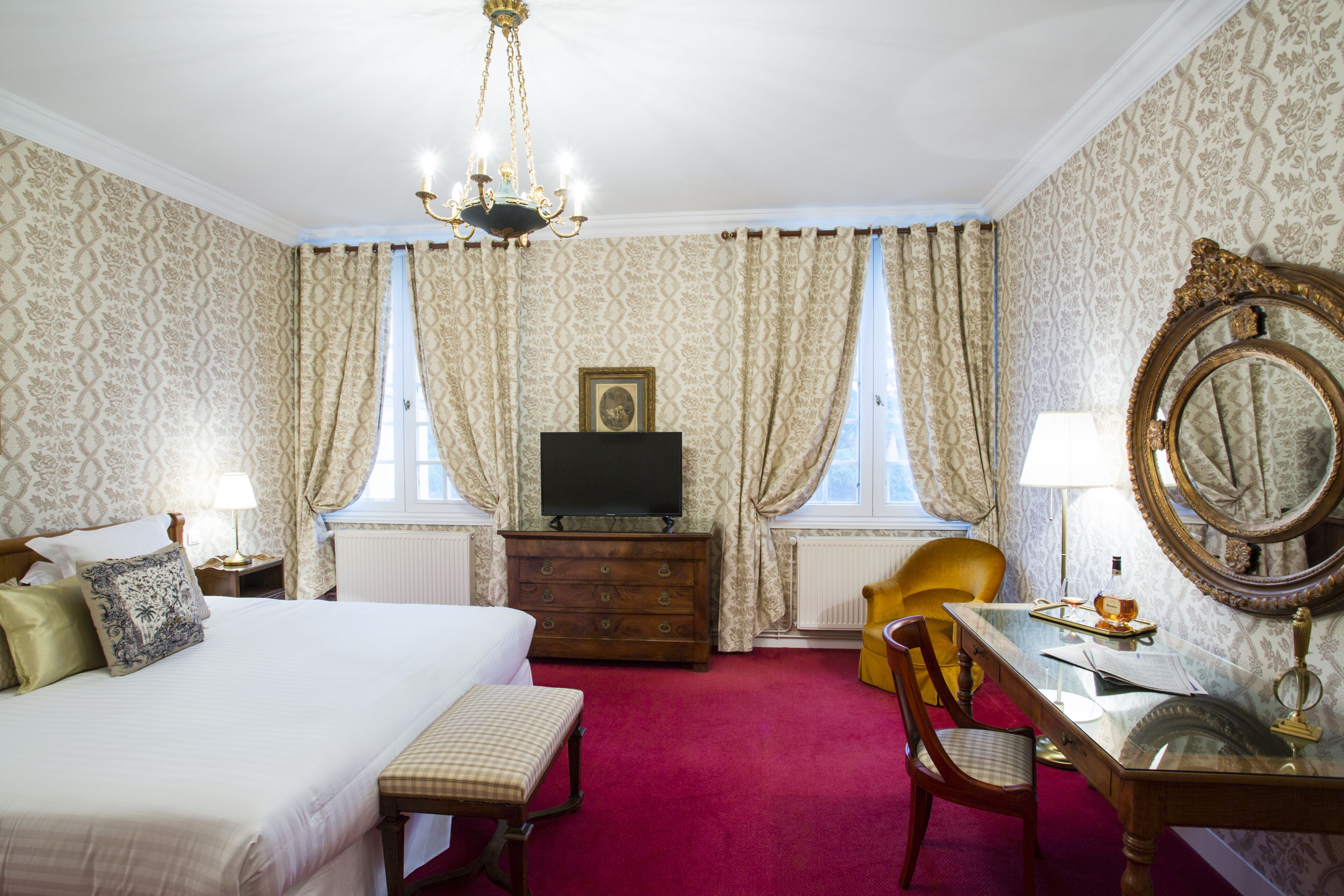 Une chambre d’hôtel avec un lit, une commode en bois, un bureau avec un miroir et une moquette rouge.