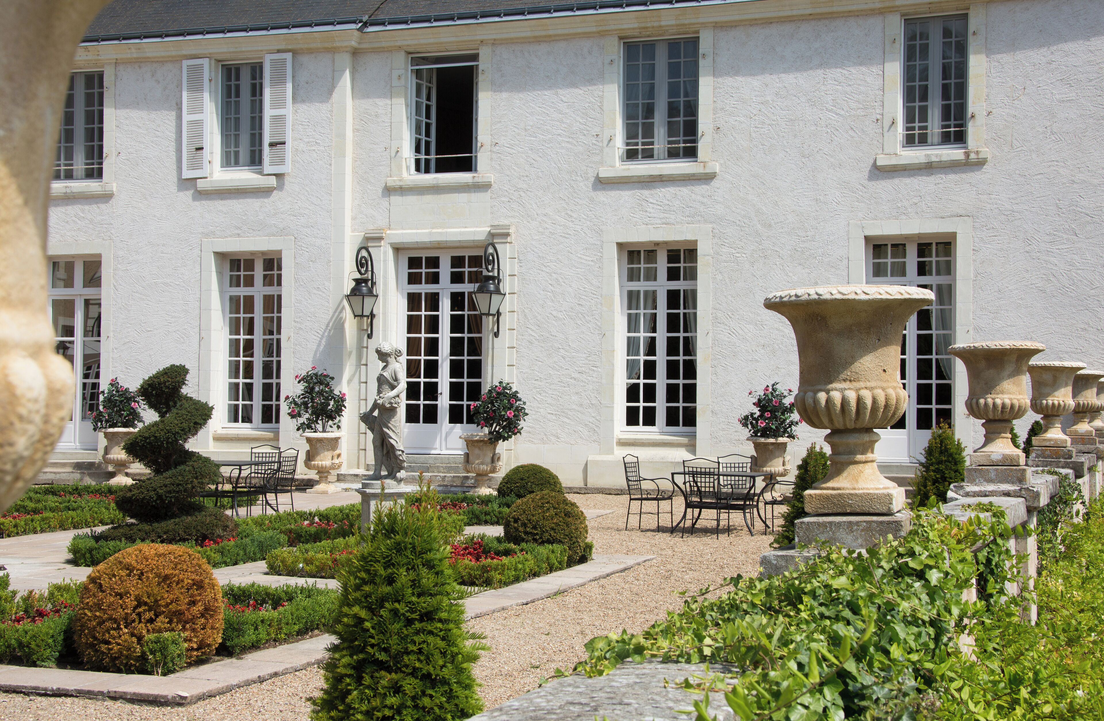 A white building with multiple windows and a garden with potted plants and outdoor furniture.