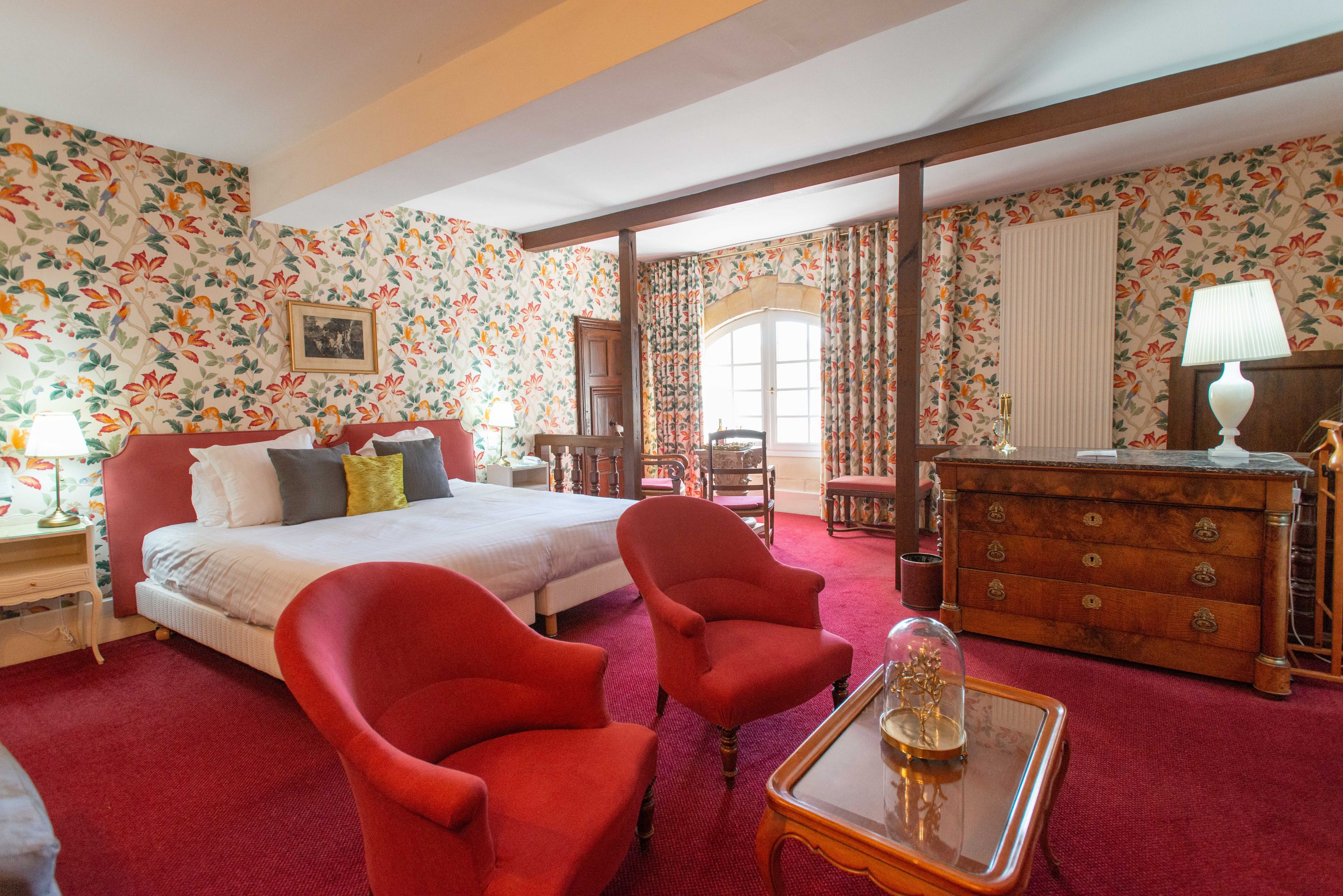A hotel room with a large bed, two red armchairs, a wooden dresser, and floral wallpaper.