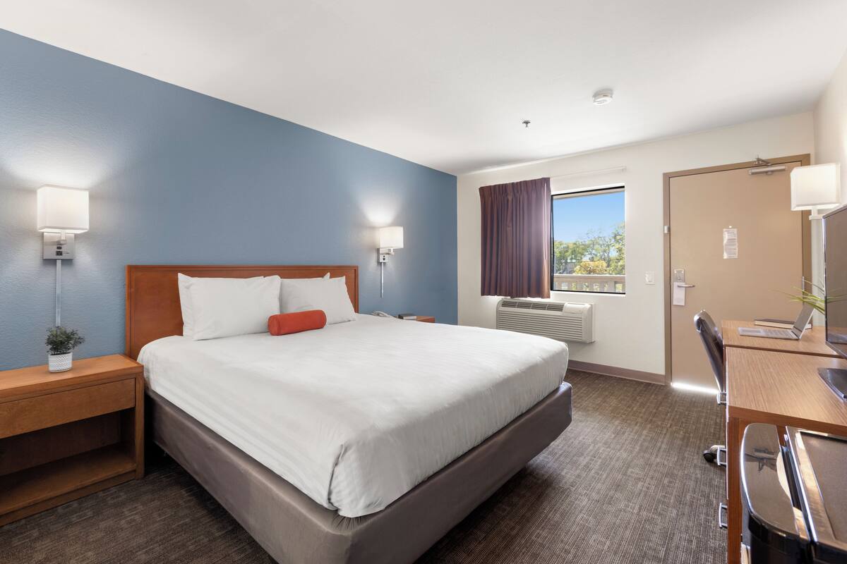 A hotel room with a bed, bedside tables, a desk with a chair, a window with curtains, and a door.