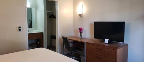 A hotel room with a bed, a desk with a chair, a television, a wardrobe, a mirror, and a wall-mounted light fixture.