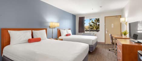 A hotel room with two beds, a desk with a computer, a TV, and a window with a view of trees.