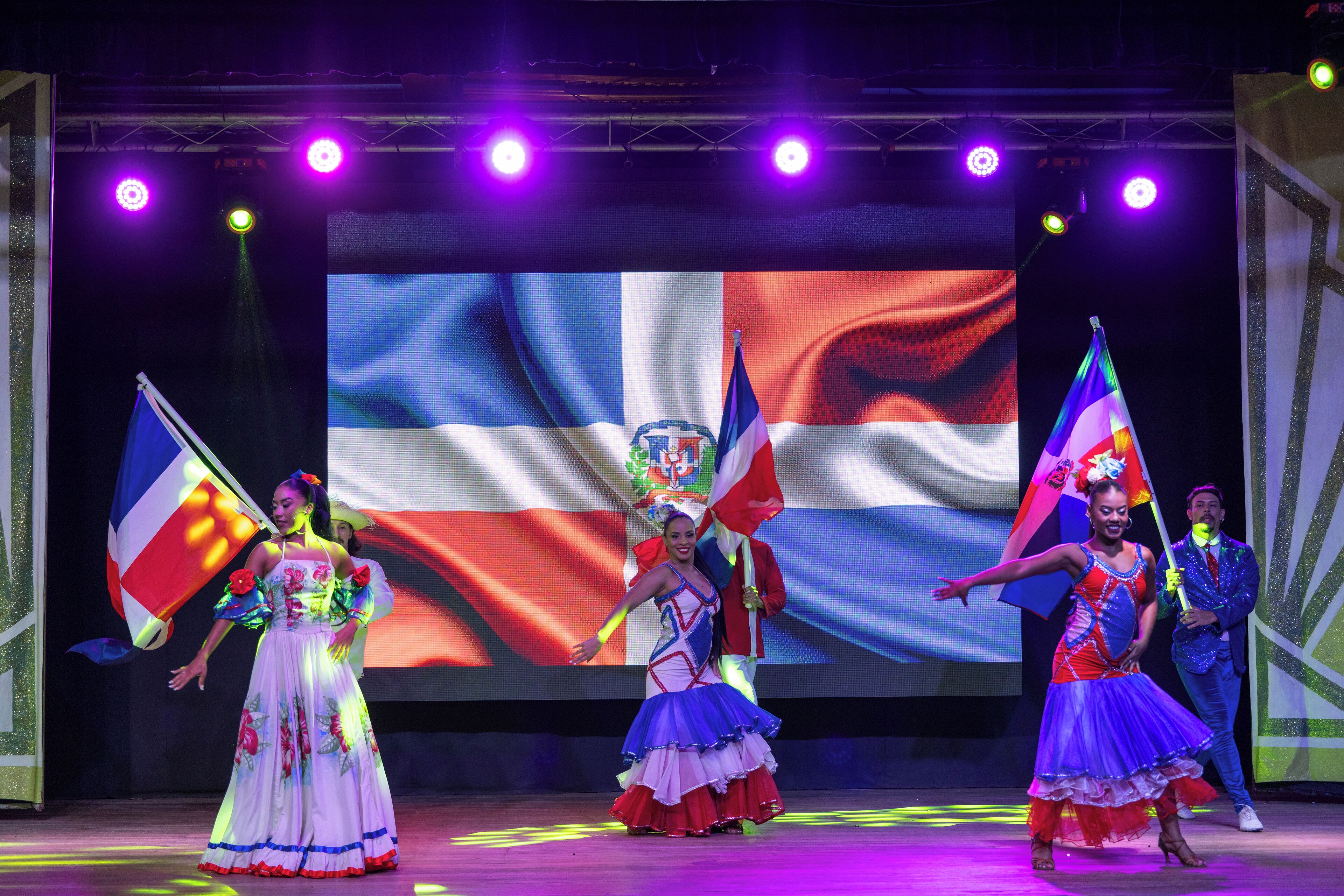 A stage with performers in colorful costumes and flags, a large screen in the background, and stage lighting.