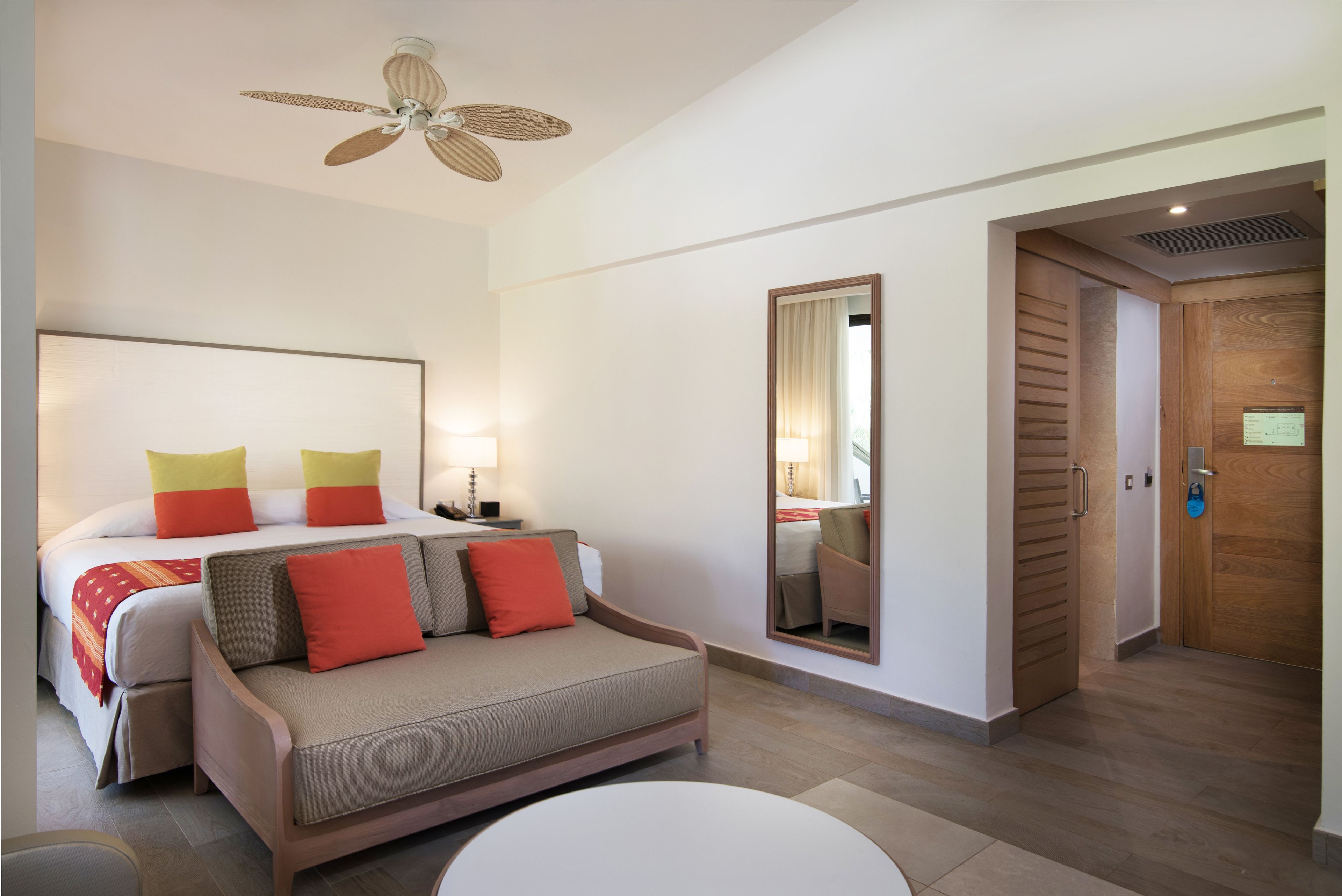 Una habitación de hotel con cama, sofá, mesa redonda, ventilador de techo, espejo y puerta de madera.
