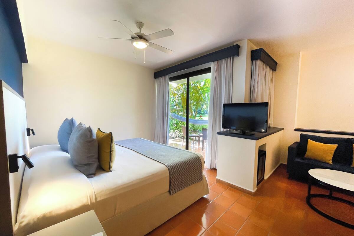 Ein Hotelzimmer mit Bett, Schreibtisch mit Fernseher, Sofa und Balkon mit Vorhängen.