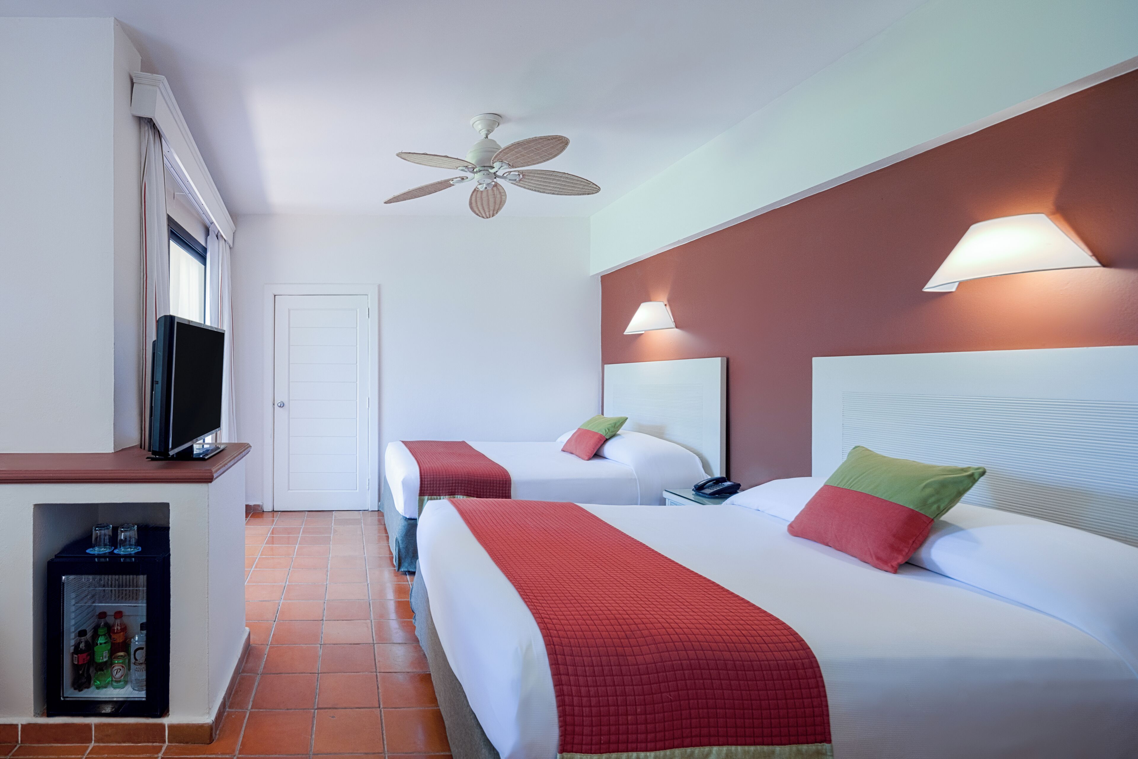 Habitación de hotel con dos camas, televisión, ventilador de techo y minibar.