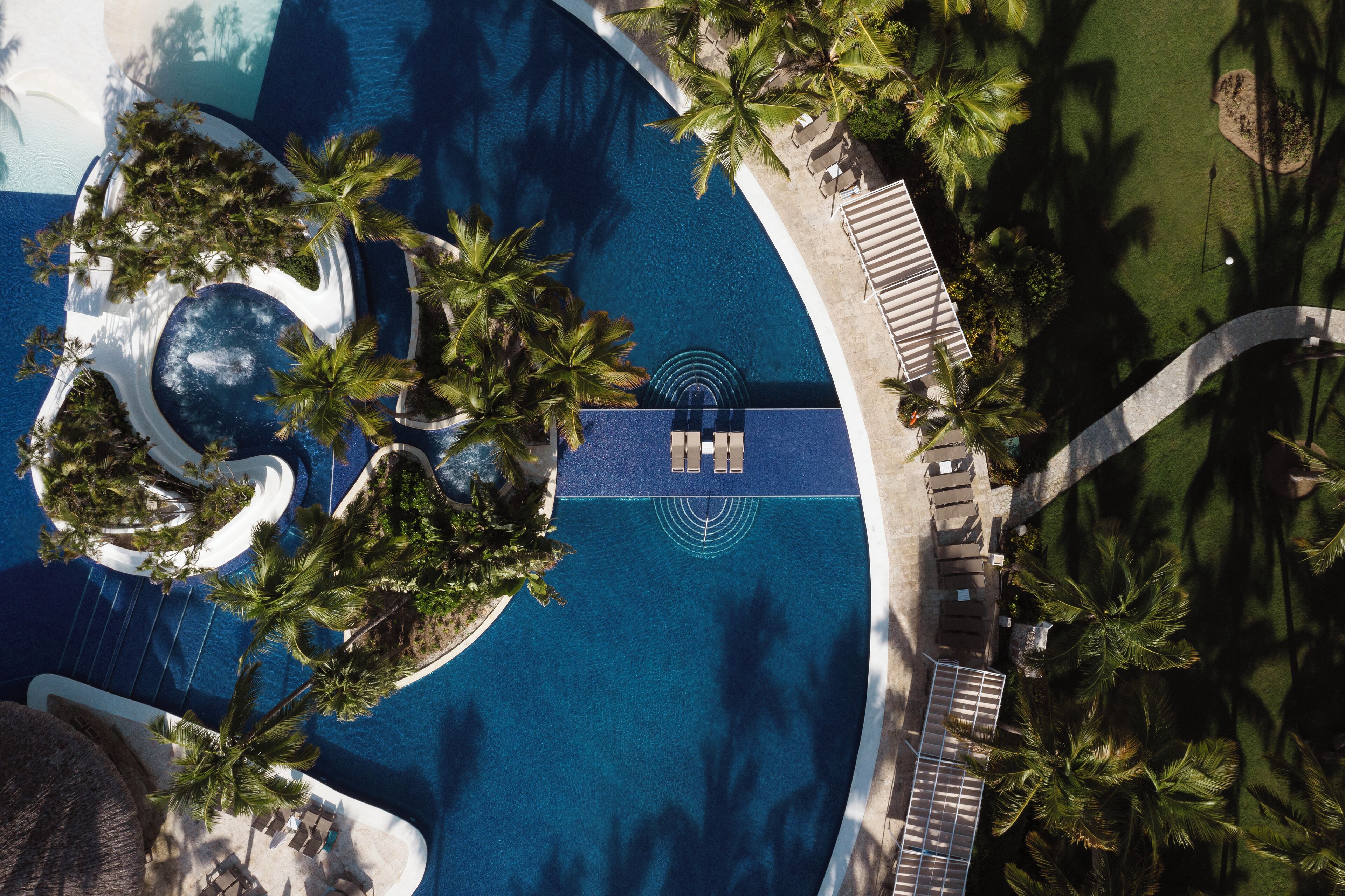 Vue aérienne d’une piscine d’un complexe hôtelier, avec des palmiers, des chaises longues et une fontaine circulaire.