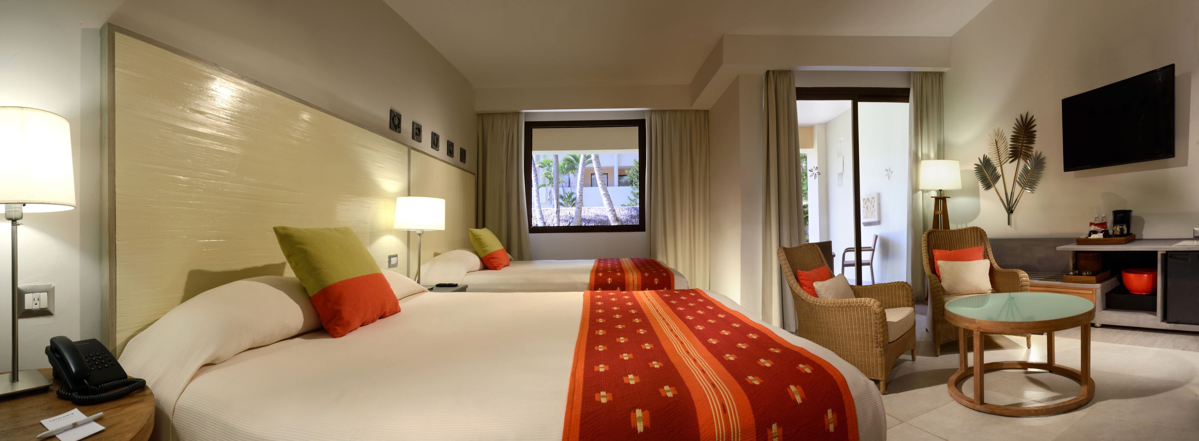 Une chambre d’hôtel avec deux lits, un coin salon comprenant une chaise et une table, une télévision et une vue sur les palmiers par la fenêtre.