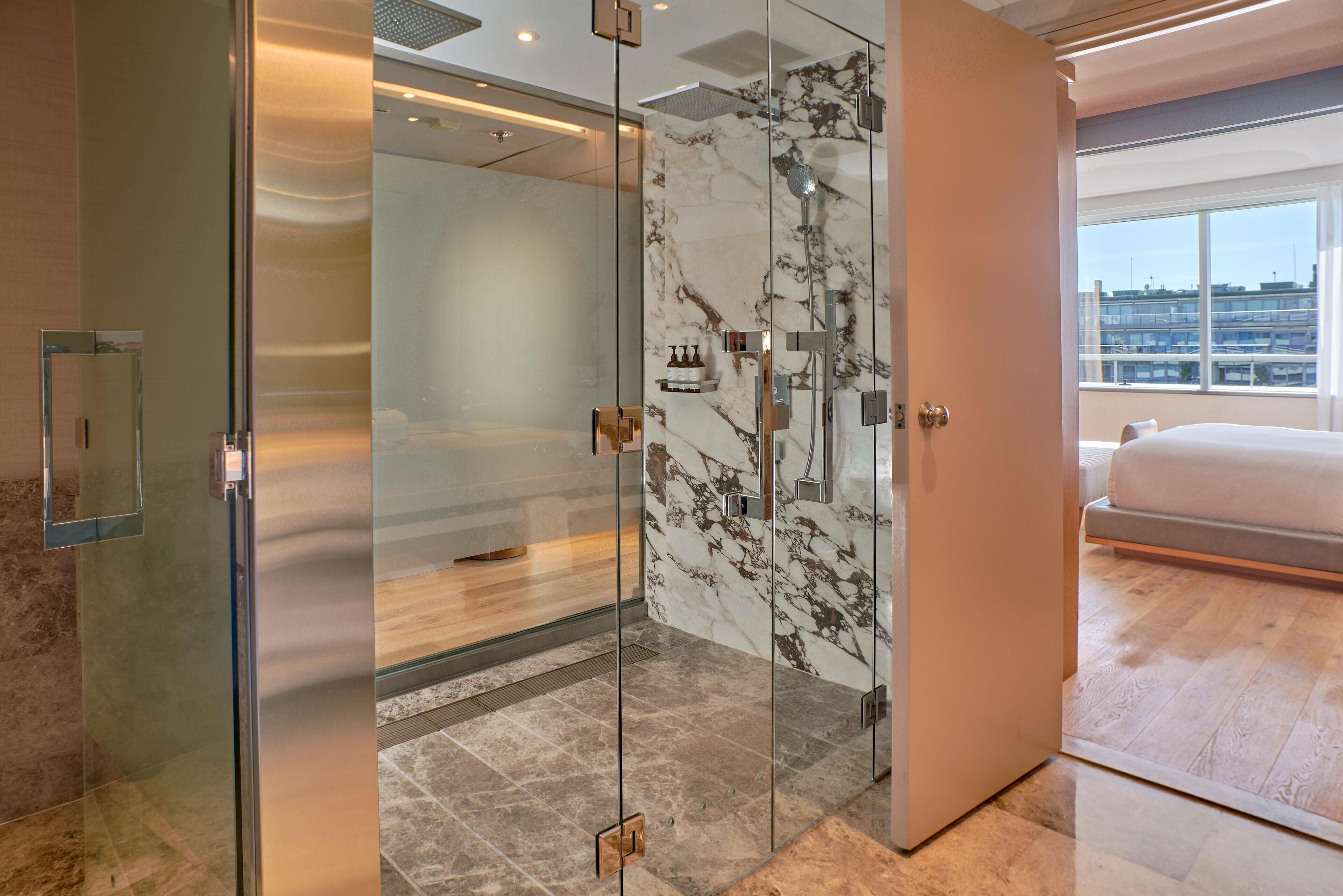 A modern hotel room with a glass-enclosed shower, marble walls, a large bed, and a city view.
