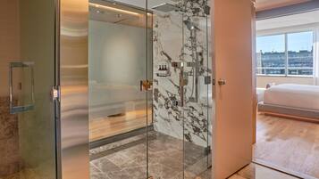 A modern hotel room with a glass-enclosed shower, marble walls, a large bed, and a city view.
