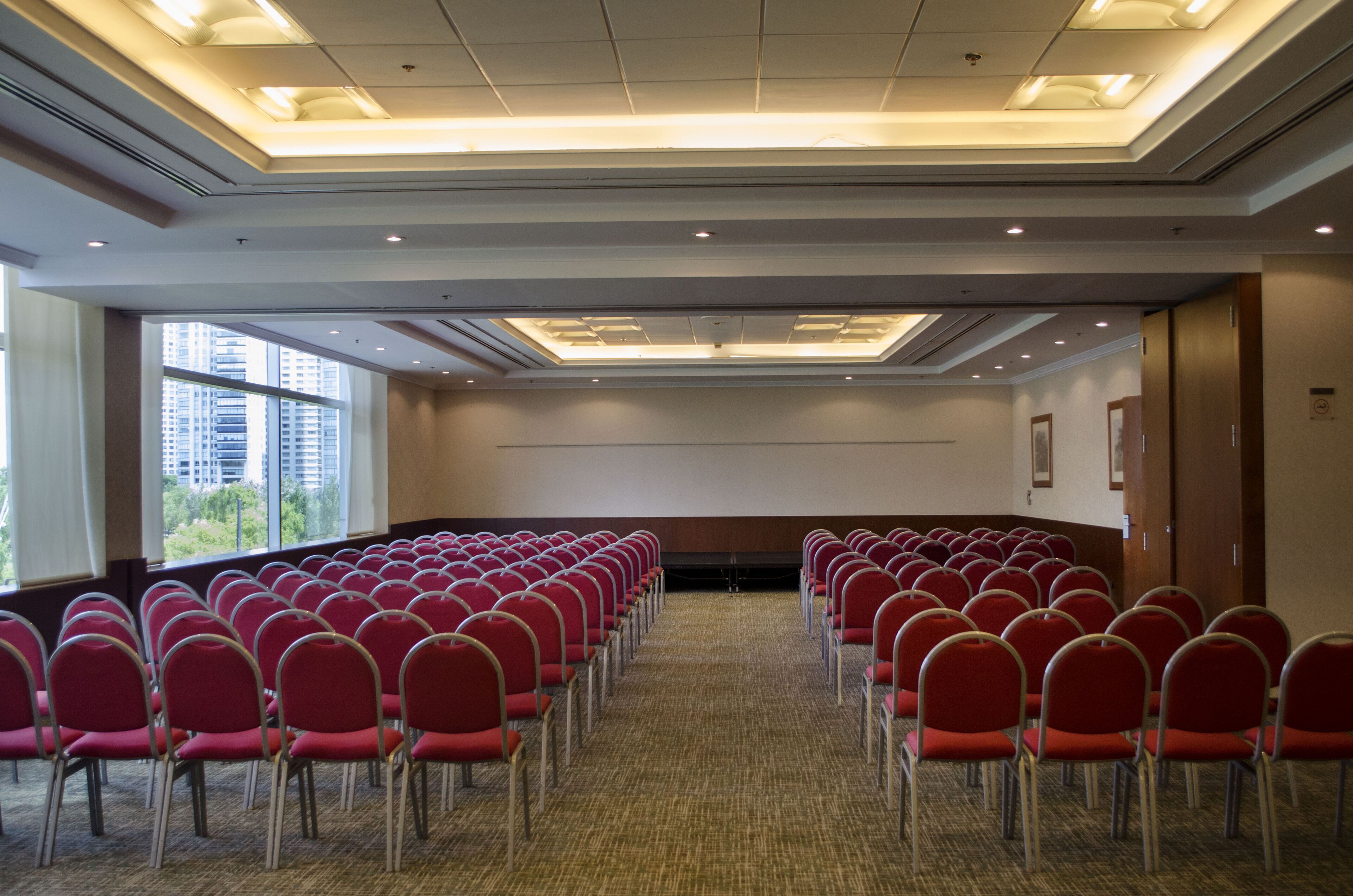 A conference room with rows of red chairs, large windows, and a carpeted floor.