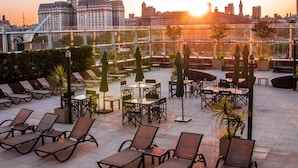 Terraza en la azotea con sillones de sol, mesas y sombrillas, con vistas a un paisaje urbano al atardecer.