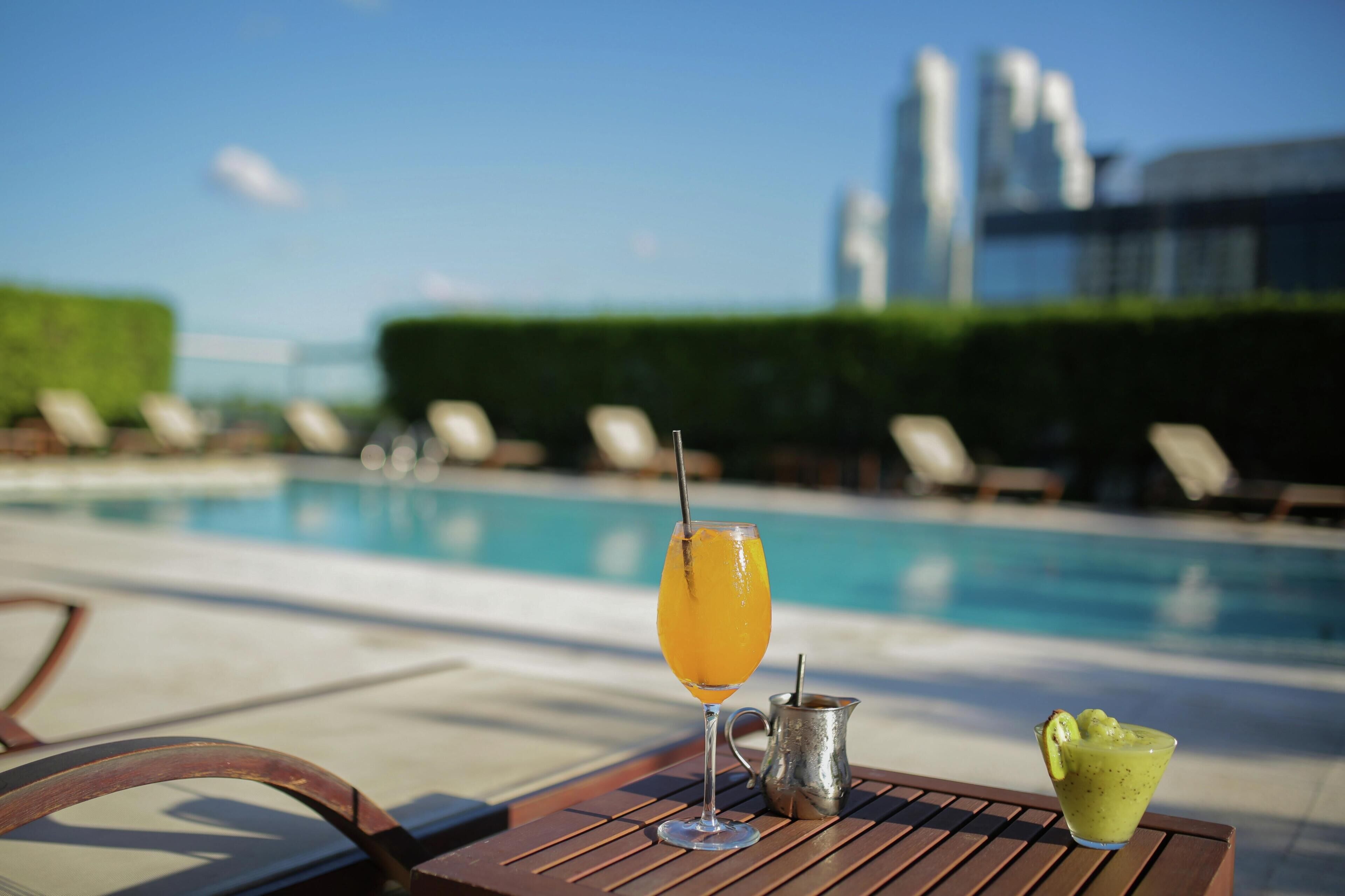 Escena junto a la piscina con un vaso de jugo de naranja, una jarra de metal y un cuenco pequeño con comida verde sobre una mesa de madera.