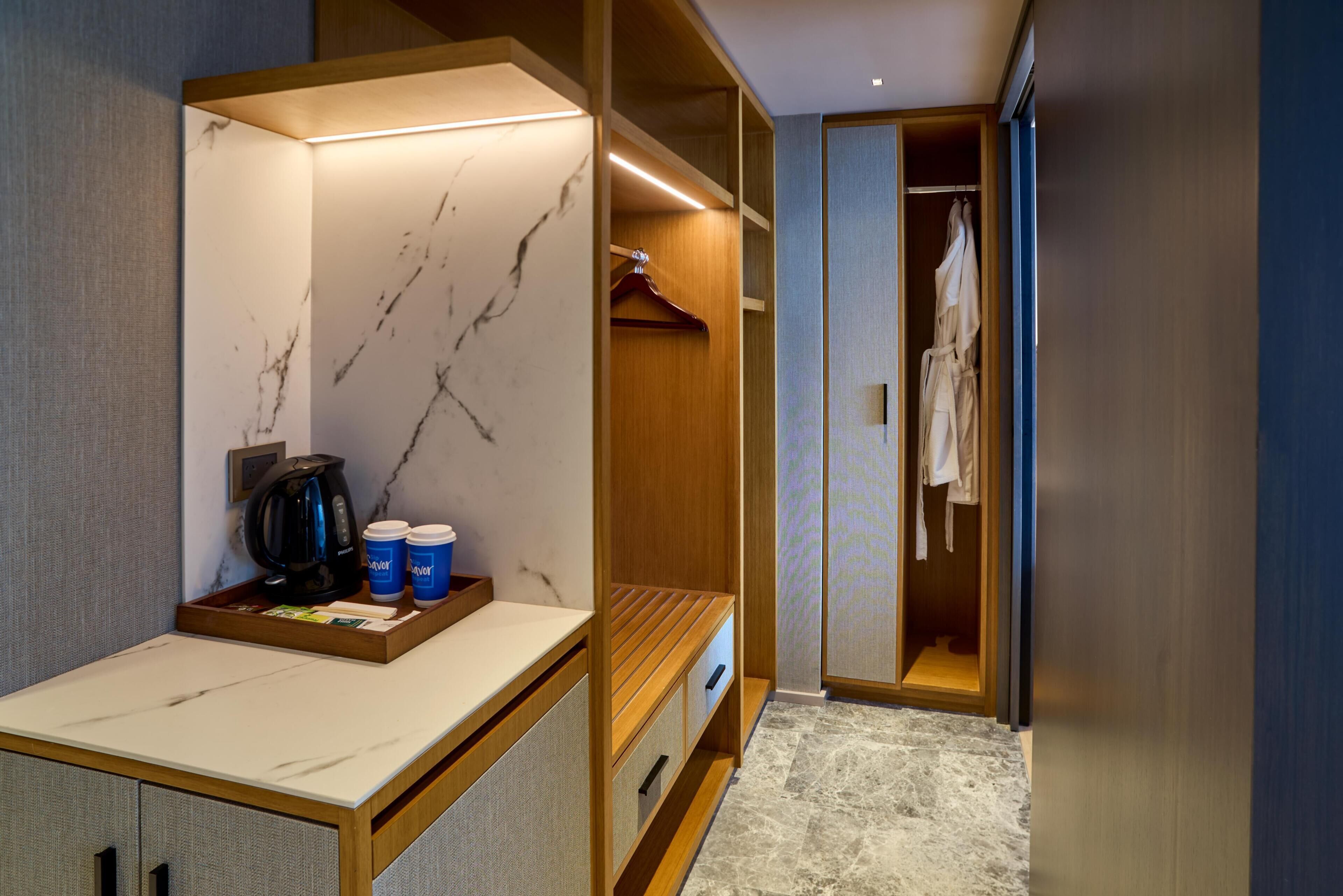 A modern hotel room with a marble wall, wooden furniture, a robe hanging, and a tray with a coffee maker and cups.