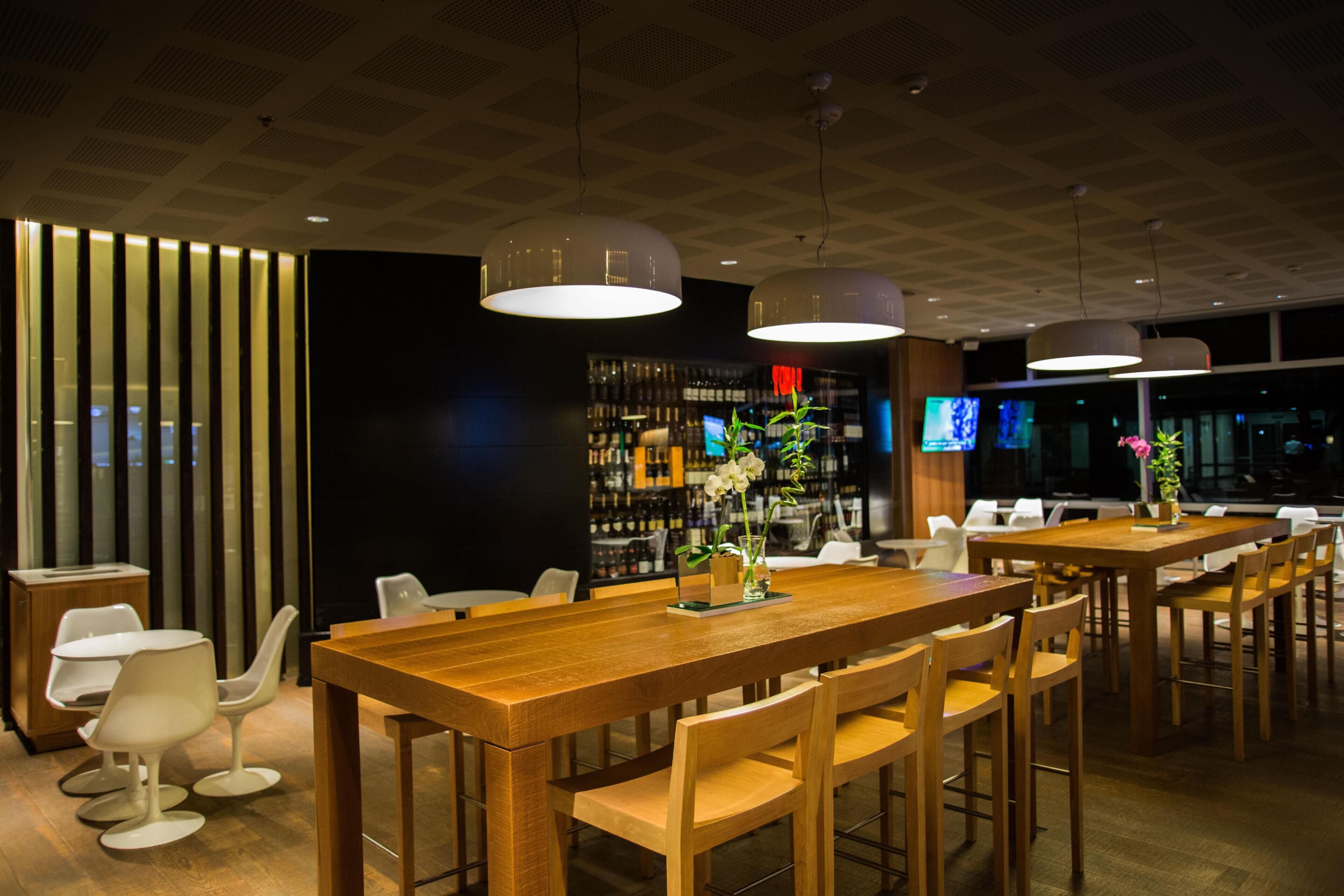 En modern restaurang med träbord och stolar, hängande lampor och en bardisk med hyllor fulla av flaskor.
