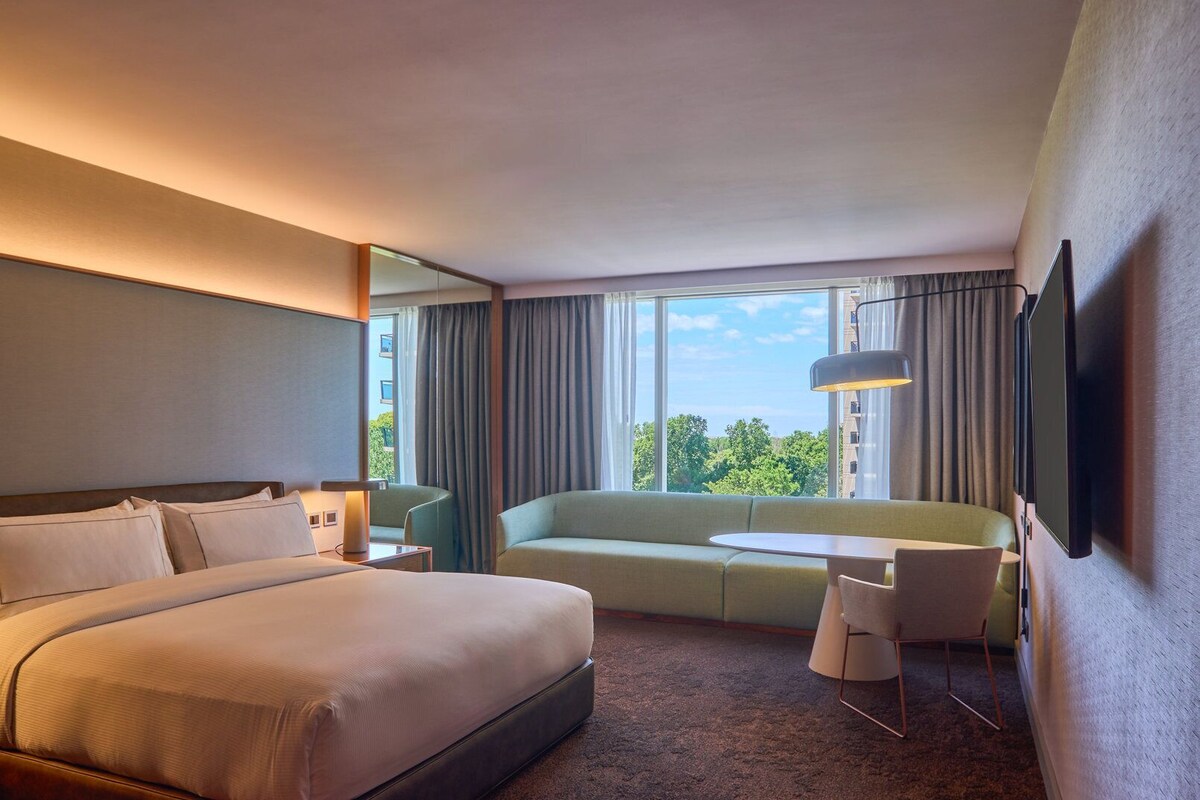 Una habitación de hotel con cama, sofá, una mesita, televisor y ventana con cortinas.