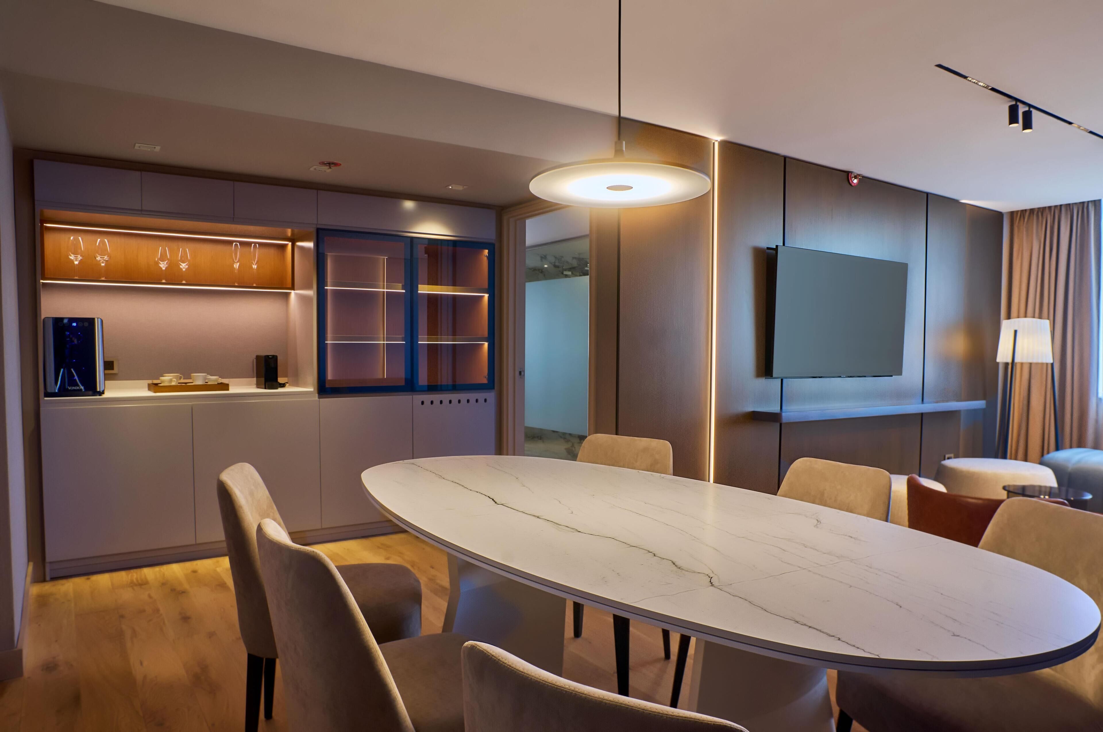 Una habitación de hotel moderna con zona de comedor, televisor de pantalla plana y un amplio ventanal.