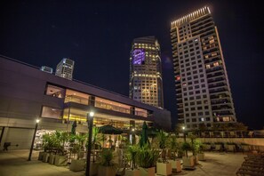 Un bâtiment moderne à plusieurs étages, doté d’un espace extérieur avec des tables et des parasols, et d’une façade bien éclairée.